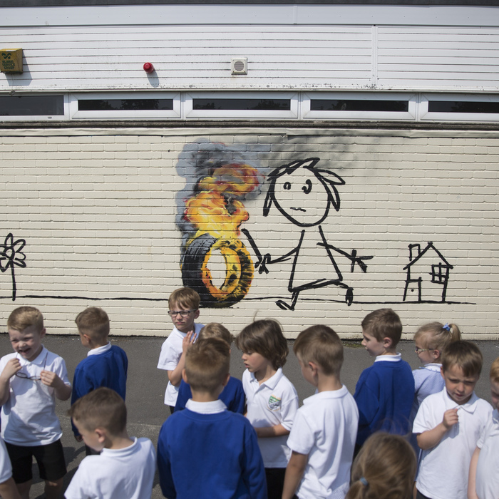 Students at the Bridge Farm Primary School in Hartcliffe, Bristol, gather round anew Banksy mural drawn recently inside the school property.  June 6 2016.  See SWNS story SWBANKSY: Pupils and teachers returned to their school after half-term to find a genuine BANKSY on the wall of classroom. The famous artist spray painted a stick-wielding child chasing a burning tyre on the side of Bridge Farm Primary School. It is believed the work is a modern take on hoop rolling, a popular game played by children during the Victorian days. The six-foot high artwork also features a flower and a small house with 'Banksy' signed to the bottom left of the brick building.